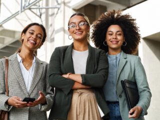 Três mulheres empresárias de blazer sorriem em frente a um prédio comercial