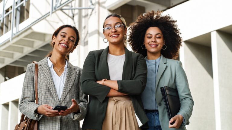 Três mulheres empresárias de blazer sorriem em frente a um prédio comercial
