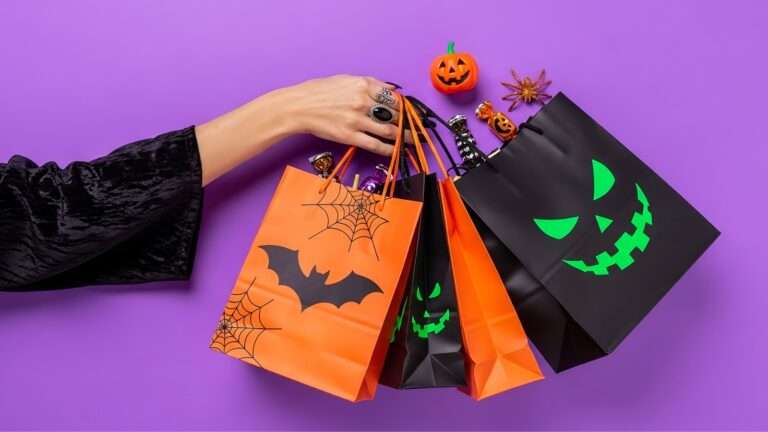 Mão segura sacolas de papel laranja e preta com estampas de Halloween, ao lado de abóbora, aranha e doces sobre fundo roxo