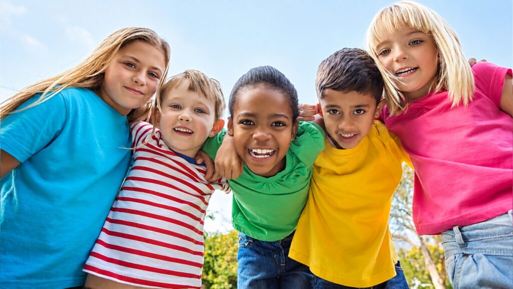 Cinco crianças sorridentes, de camisetas coloridas, abraçadas ao ar livre sob céu azul