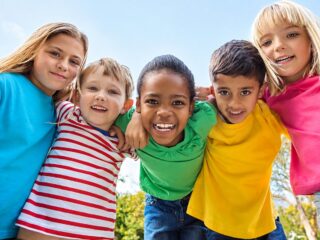 Cinco crianças sorridentes, de camisetas coloridas, abraçadas ao ar livre sob céu azul